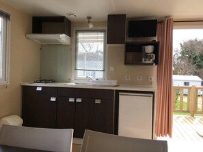 A compact kitchenette with a stove, sink, and a small refrigerator. There is a window with blinds, a ceiling fan, and a view of a deck outside. - Camping 3 étoiles - Piscine  - ccbgchf (Perros-Guirec)