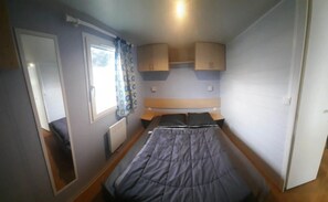 A compact room with a bed, a window with curtains, a radiator, and wooden cabinets. - Camping 3 étoiles - Piscine  - ccbgcib (Perros-Guirec)