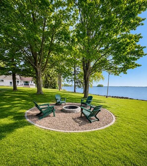 A circular seating area with chairs and a fire pit surrounded by a gravel ring, set in a grassy field with trees and a lake view.