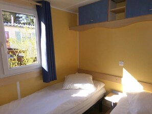 A small, single-bed room with a window, a curtain, a bedside table, and a blue cabinet. - Camping 3 étoiles - ccbgdg0 (Neuwiller-lès-Saverne)