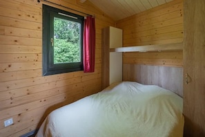 A single bed in a cabin with wooden walls, a window with a curtain, and a small white cabinet.