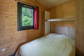 A single bed in a cabin with wooden walls, a window with a curtain, and a small white cabinet.