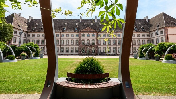 A courtyard with a central bench, surrounded by greenery and a large building with multiple windows and a symmetrical facade. - Le Nid d'Alspach: Romantic Retreat in Colmar (Colmar)