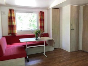 A room with a red sofa, a small table with a vase, a window with curtains, and a door. - Camping 3 étoiles - Piscine  - ccbgdcf (Rennes-les-Bains)