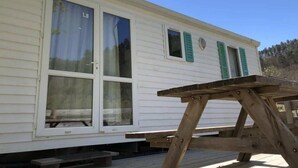 Una casa móvil blanca con contraventanas verdes, una terraza de madera y una mesa de picnic.