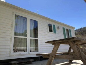 A white mobile home with green shutters, a wooden deck, and a picnic table. - Camping 3 étoiles - Piscine  - ccbgdcf (Rennes-les-Bains)