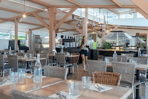 Ein Restaurant im Freien mit Holztischen und Korbstühlen, einem Küchenbereich im Hintergrund und freiem Blick auf den Himmel.