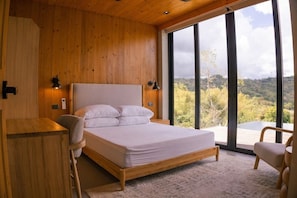 A bedroom with a wooden bed, a desk, and a chair, large windows with a view of trees and hills.