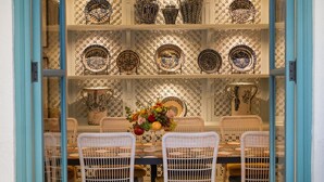 Zona pranzo con tavolo apparecchiato per quattro, circondato da sedie in vimini bianco, un lampadario e una parete di piatti decorativi.
