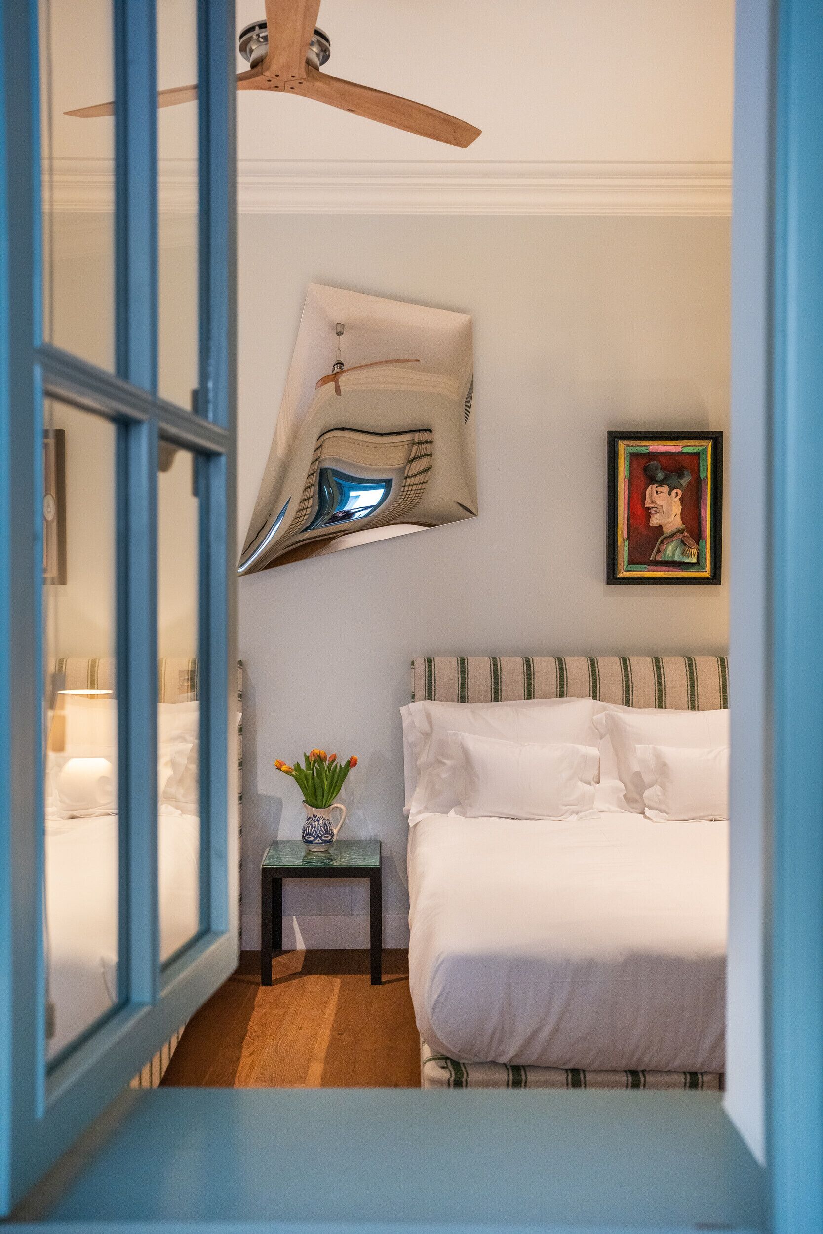 A bedroom with a bed, a small table with a vase, a ceiling fan, and a framed picture on the wall.