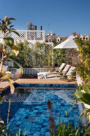 Ein Poolbereich mit Liegestühlen, einer gefliesten Terrasse und Blick auf Gebäude im Hintergrund.