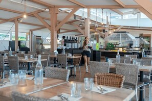 Ein Restaurant im Freien mit Holztischen und Korbstühlen, einem Küchenbereich im Hintergrund und freiem Blick auf den Himmel.