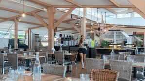 Ein Restaurant im Freien mit Holztischen und Korbstühlen, einem Küchenbereich im Hintergrund und freiem Blick auf den Himmel.