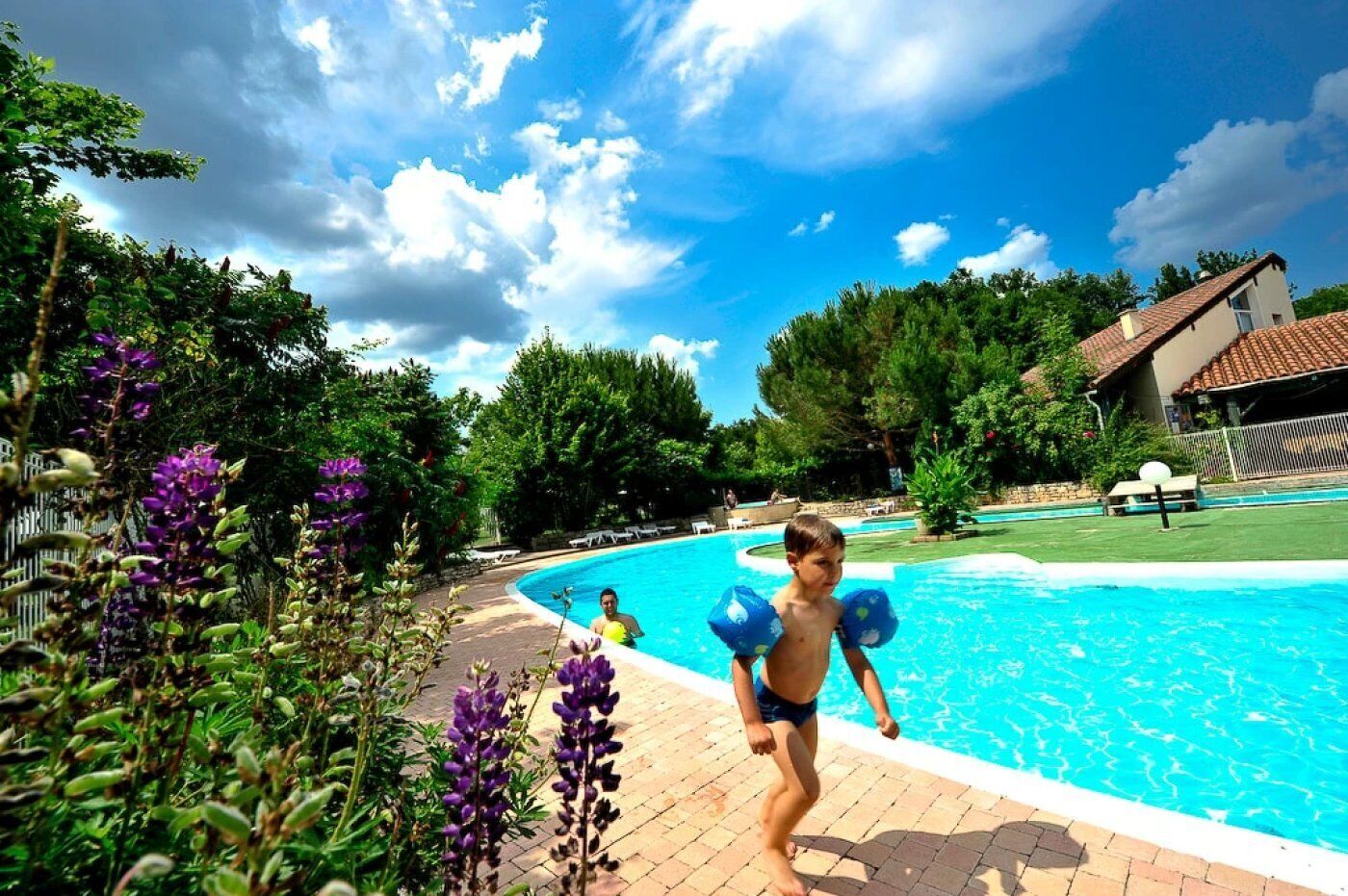 Une piscine sous un ciel dégagé, entourée de verdure et bordée d’un bâtiment dont le toit est recouvert de tuiles.