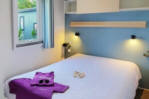 Ein Einzelbett mit weißer Bettdecke, einem lila Hemd und einer Sonnenbrille. Ein Fenster mit Vorhängen, ein wandmontiertes Regal und eine Wandleuchte sind vorhanden.