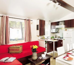 A compact living space with a red sofa, a small dining table, a kitchen area with a microwave and stove, and a window with curtains. - Camping 4 étoiles - Parc aquatique - ccbgdeb (La Baule)