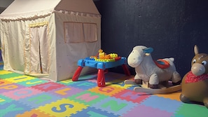 Ein Spielbereich mit Zelt, Spieltisch mit bunten Bauklötzen und zwei Stofftieren auf einer farbenfrohen Spielmatte.