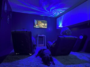 A room with a large screen, two chairs, a small table, and a dog on a rug.