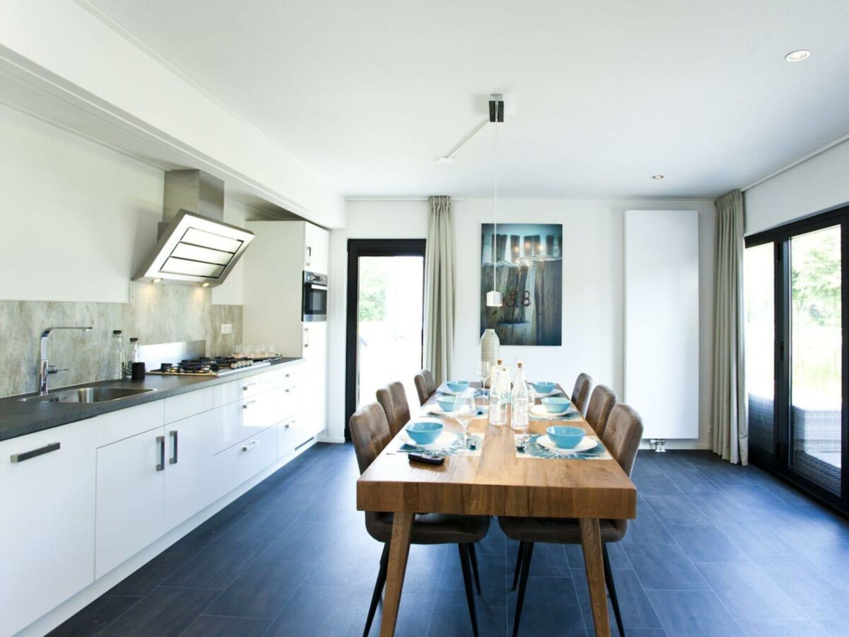 A modern kitchen with a dining area, featuring a large table, chairs, and a view through the glass door.