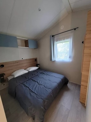 Ein Schlafzimmer mit Bett, einem Fenster mit Vorhängen, einem hölzernen Kopfteil und einem kleinen Regal.