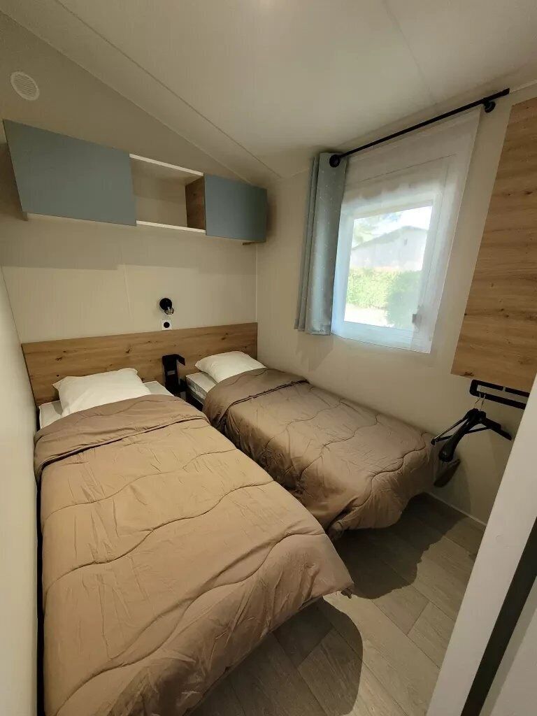 Een kleine slaapkamer met twee bedden, een raam met gordijn, een houten hoofdbord en een wandplank.