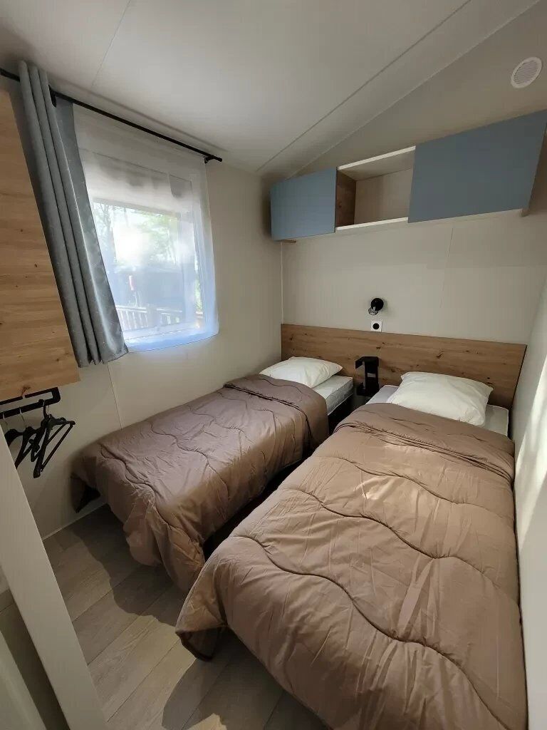 A small, compact room with two single beds, a window with curtains, a wooden headboard, and a wall-mounted shelf.