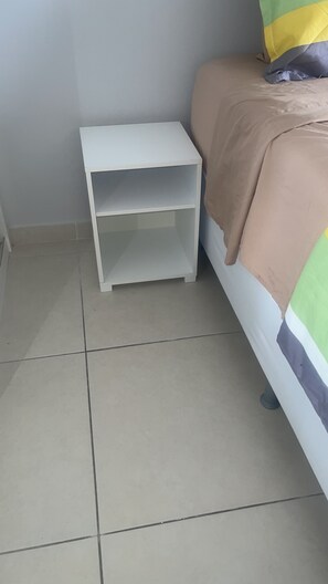 Quarto com uma cama, mesa de cabeceira branca e piso de azulejos.