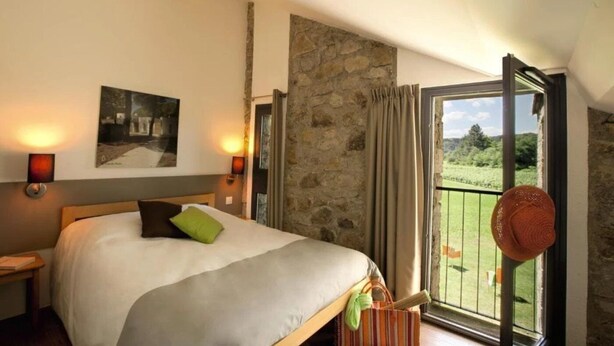 A bedroom with a bed, two wall-mounted lamps, a stone wall, a window with curtains, and a view of greenery outside. - Camping 3 étoiles - Piscine  - ccbc0gc (Chambonas)