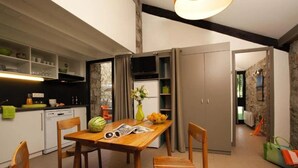 A modern kitchen with a dining area, wooden chairs, a table with a watermelon and flowers, and a stone wall. - Camping 3 étoiles - Piscine  - ccbc0gc (Chambonas)