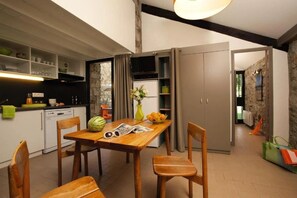 A modern kitchen with a dining area, wooden chairs, a table with a watermelon and flowers, and a stone wall. - Camping 3 étoiles - Piscine  - ccbc0gc (Chambonas)