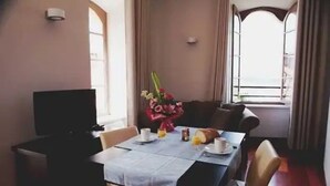 Uma sala de jantar com mesa posta para dois, uma televisão em um suporte e uma janela com cortinas.