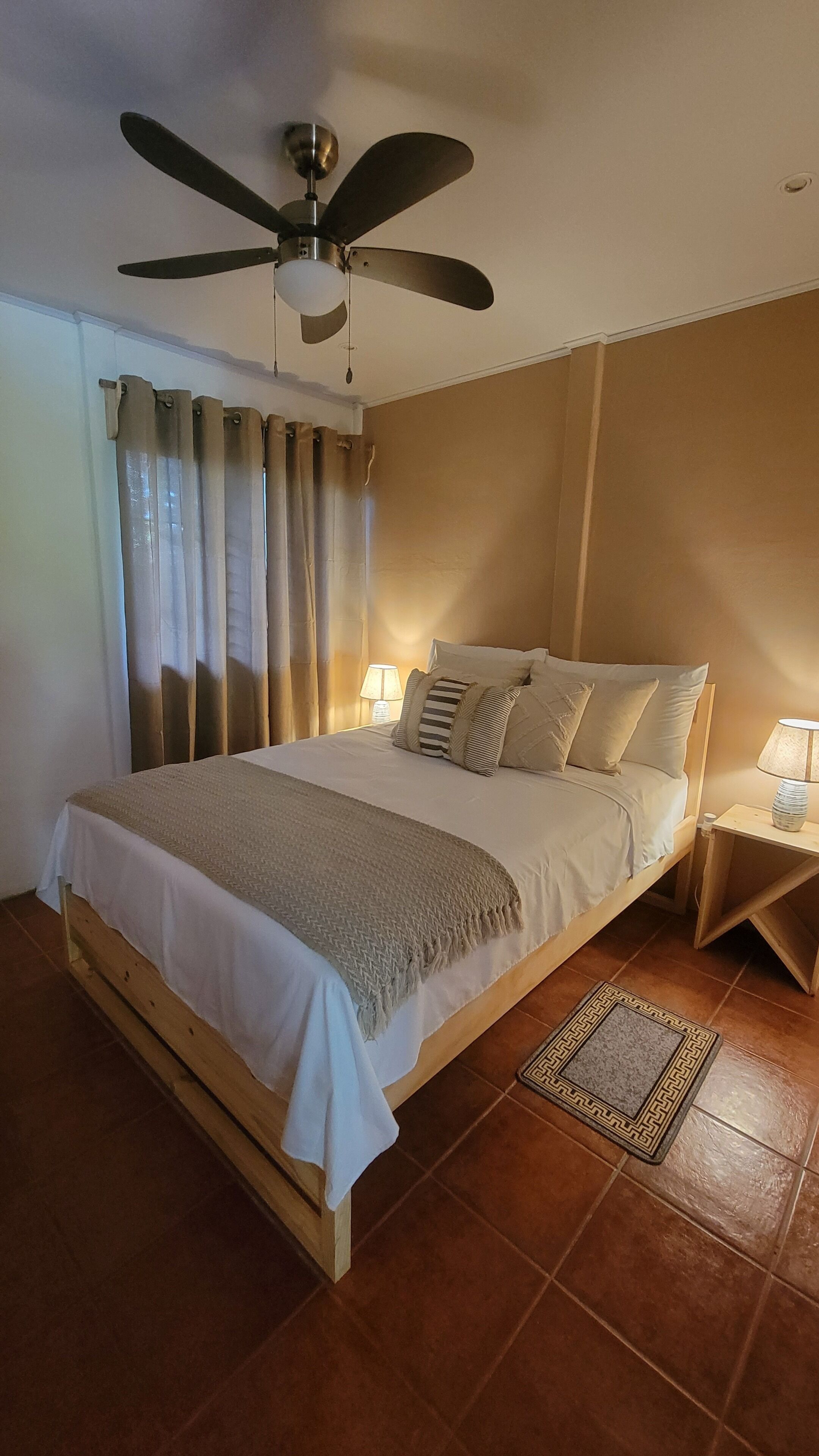 Une chambre à coucher avec un lit, des rideaux, un ventilateur de plafond, des lampes de chevet et un tapis.