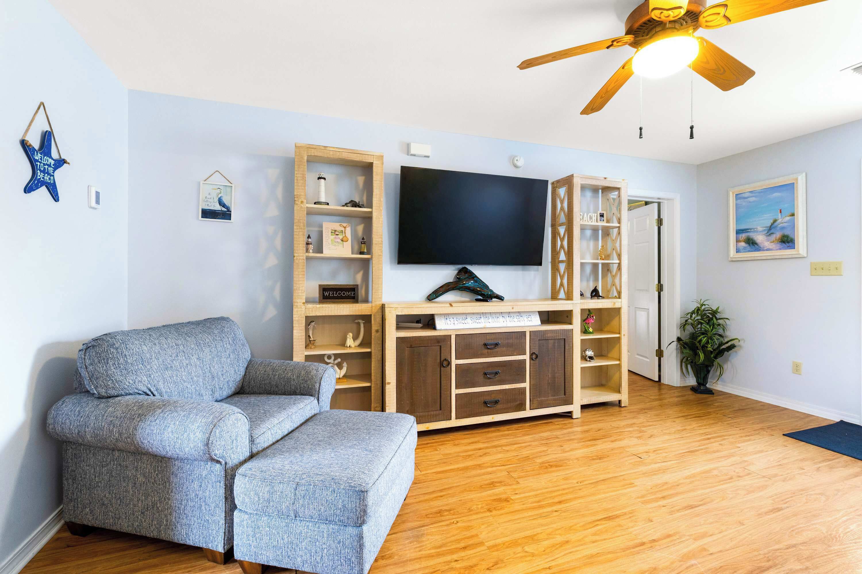 Uma sala de estar com sofá cinza, unidade de entretenimento de madeira, televisão de tela plana, ventilador de teto e um vaso de planta.