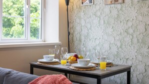 Una mesa de desayuno con platos, tazones, vasos y fruta, frente a un papel tapiz floral y una ventana con vistas a árboles.