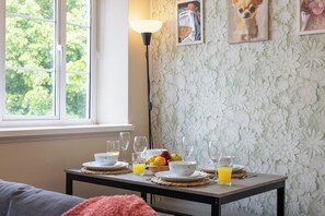 Ett frukostbord med tallrikar, skålar, glas och frukt, placerat framför en blommig tapet och ett fönster med utsikt över träd.