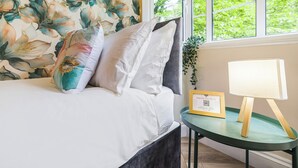 Una cama con sábanas blancas, un papel tapiz floral, una mesita de noche con lámpara y un certificado enmarcado.