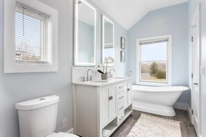 A bathroom with a toilet, a vanity with a mirror, a bathtub, and a window with blinds. - Waterfront Retreat w/ Dock & Modern Design (Center Moriches)