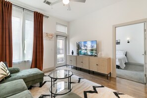 Ein modernes Wohnzimmer mit einer Couch, einem Glastisch, einem Fernseher auf einer Kommode und einer Tür zu einem Schlafzimmer.