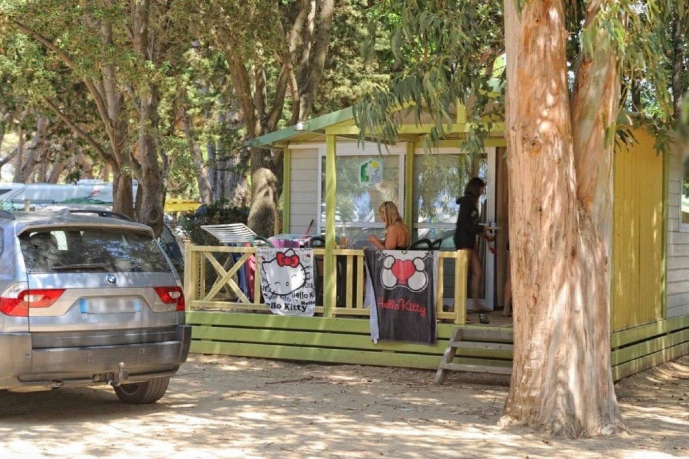 A small outdoor café with a green and yellow structure, a car parked nearby, and trees in the background.