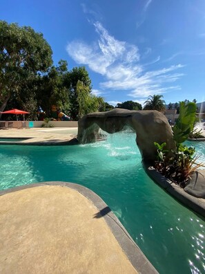 A pool with a rock formation, clear blue water, and surrounding greenery.