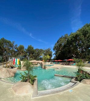A pool area with multiple water features, including a central pool and smaller water channels, surrounded by trees and shaded by umbrellas.