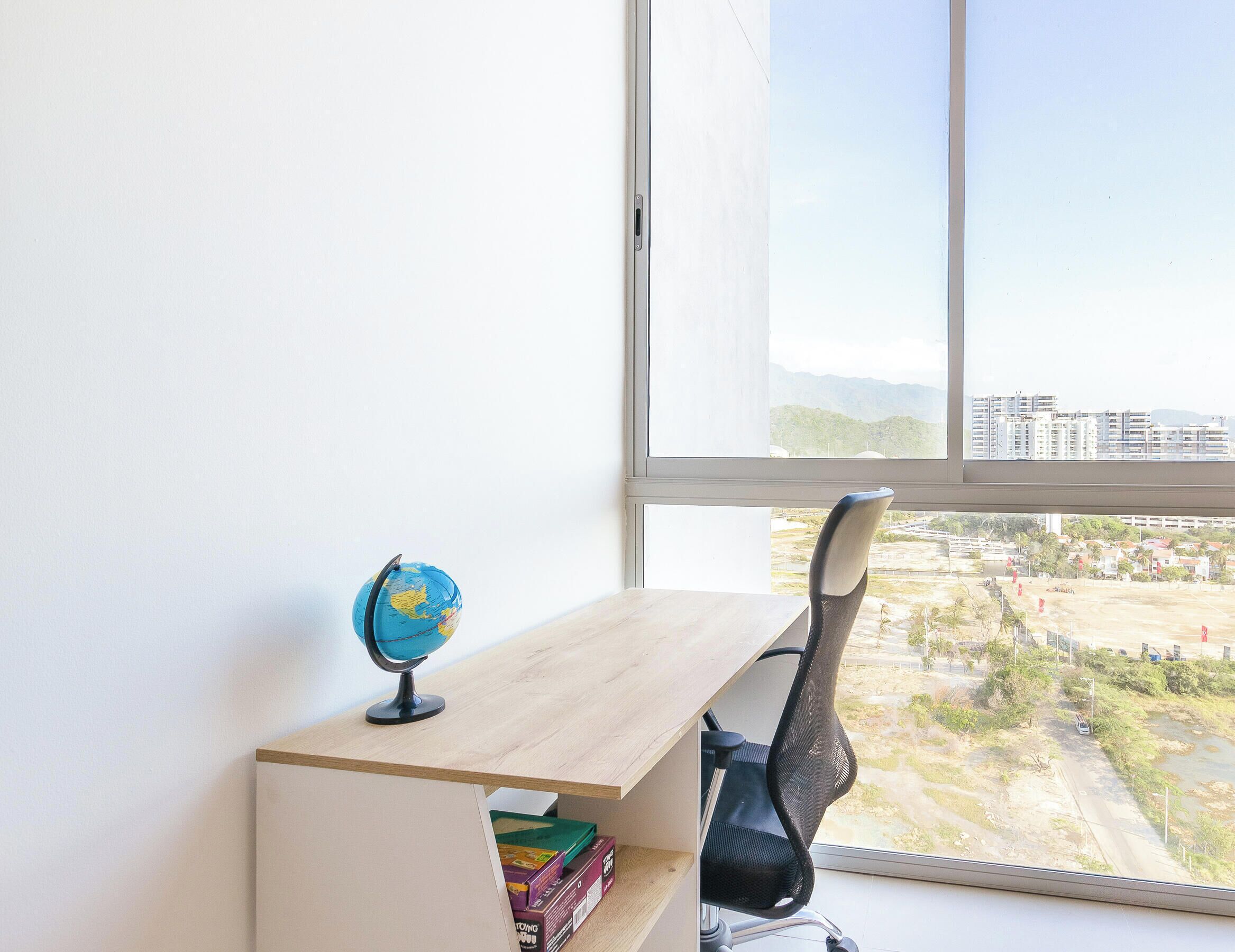 Quarto com escrivaninha, cadeira, globo e janela com vista para edifícios e estrada.