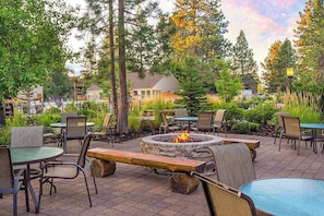 An outdoor patio area with fire pit seating, tables, and chairs, surrounded by trees and a view of houses in the background. - 1BR Condo, Pool & Hot Tub, fitness center, trails (Bend)