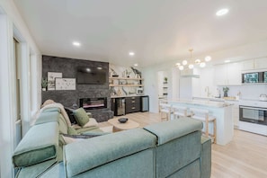 A modern living room with a sofa, a fireplace, and a kitchen area with white cabinets and a dining space. - 1BR Condo, Pool & Hot Tub, fitness center, trails (Bend)