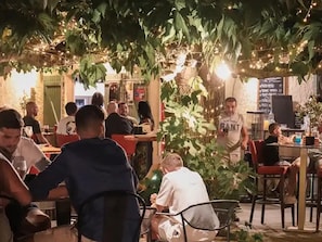 Un espace de restauration en plein air, avec des tables et des chaises, entouré de verdure et illuminé de guirlandes lumineuses.