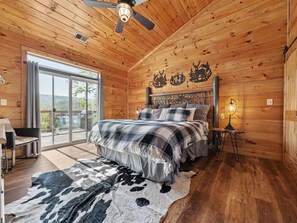 Ein Schlafzimmer in einer Holzhütte mit einem Bett, einem Nachttisch, einer Lampe, einem großen Fenster und Blick ins Freie.