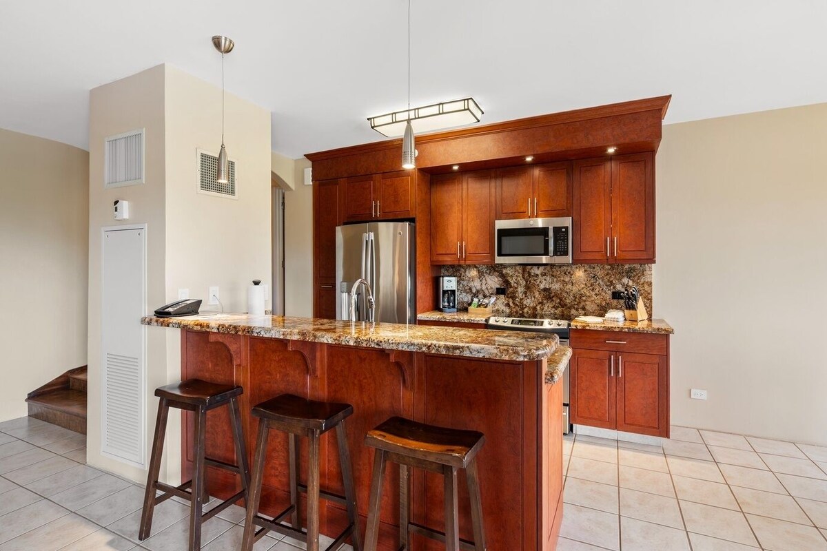 A kitchen with wooden cabinets, a granite countertop, stainless steel appliances, and a bar stool area.