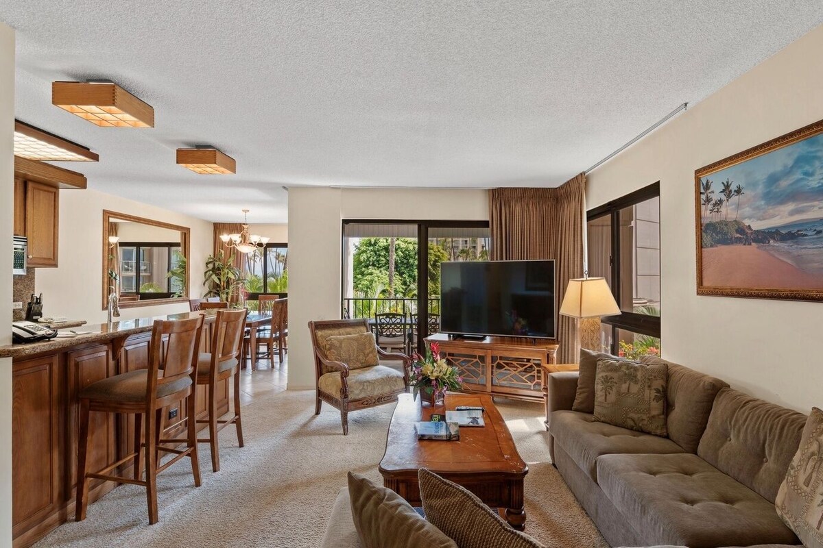 A spacious living area with a large flat-screen TV, a sofa, a coffee table, and a dining area with chairs and a kitchen visible in the background.
