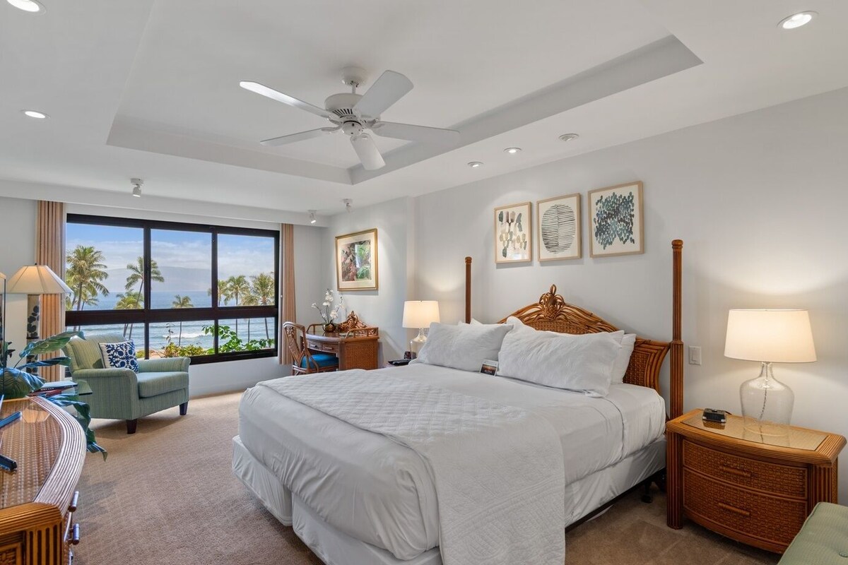 A hotel room with a large bed, a ceiling fan, a view of the ocean, and artwork on the walls.