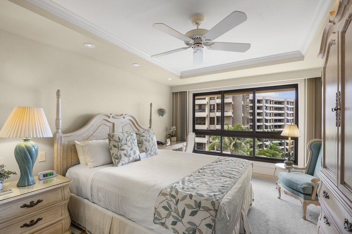 A bedroom with a bed, bedside table, lamp, clock, and a view of buildings through a large window.